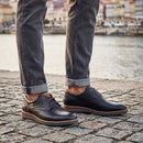 A person wearing dark gray cuffed jeans and black leather lace-up dress shoes with brown laces, standing on a cobblestone surface near a waterfront.