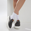 Person wearing black slip-on shoes with white socks on a light background