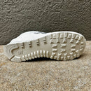 A close-up of the outsole of a white New Balance sneaker, showing the tread pattern and embossed 'NB' logo, resting on a textured concrete surface.