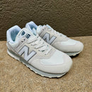 A pair of New Balance 574 sneakers in light gray and white, displayed on a textured concrete surface against a brown wall.