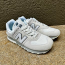 A pair of New Balance 574 sneakers in light gray and white, featuring the signature 'N' logo, lace-up closure, and light blue lining, set against a textured brown background.