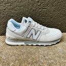 A light gray and white New Balance sneaker with a light blue lining, shown in a side profile against a textured brown wall and concrete floor.