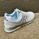 A single New Balance sneaker in white and light gray with a pale blue lining, photographed on a textured concrete surface against a stucco wall.