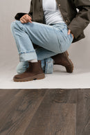 A person crouching in light blue jeans, a white top, a dark olive jacket, and brown Chelsea-style boots, photographed in a studio with a light neutral backdrop and dark wood flooring visible at the bottom.