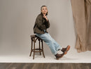 A model sits on a dark wooden stool wearing a dark brown jacket, light blue jeans, and brown leather boots in a studio setting with a beige backdrop and a draped fabric panel.