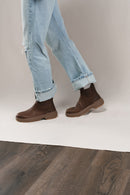 A person wearing light blue cuffed jeans and dark brown Chelsea-style ankle boots, standing on a white floor riser over a dark wood floor against a light beige studio backdrop.