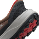Close-up of a trail running shoe showing the heel, upper, midsole, and outsole. The shoe features a black and dark gray upper with a brown collar, a thick light gray midsole, and a black and red treaded outsole.