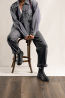 A person seated on a wooden stool wearing a faded gray denim jacket, dark gray jeans, and black rubber boots against a light neutral studio backdrop with a wood floor.