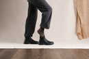 A person wearing black Chelsea-style rain boots with dark gray cuffed jeans and a brown sock, standing on a white floor against a beige wall with a wood floor visible.