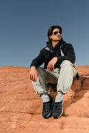 A man wearing a black hooded jacket, light gray jogger pants, gray socks, and black slip-on shoes sits on red rock formations under a clear blue sky.