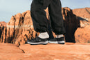 Black and white athletic shoes with white socks, worn by a person standing on a red rock desert trail with canyon cliffs in the background.